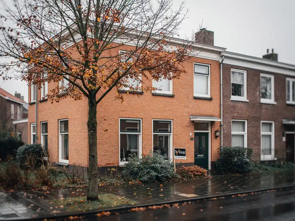 Nederlandstalige rijwoning in Waddinxveen te koop, met herfstkleuren in de boom en nat trottoir onder bewolkte lucht.