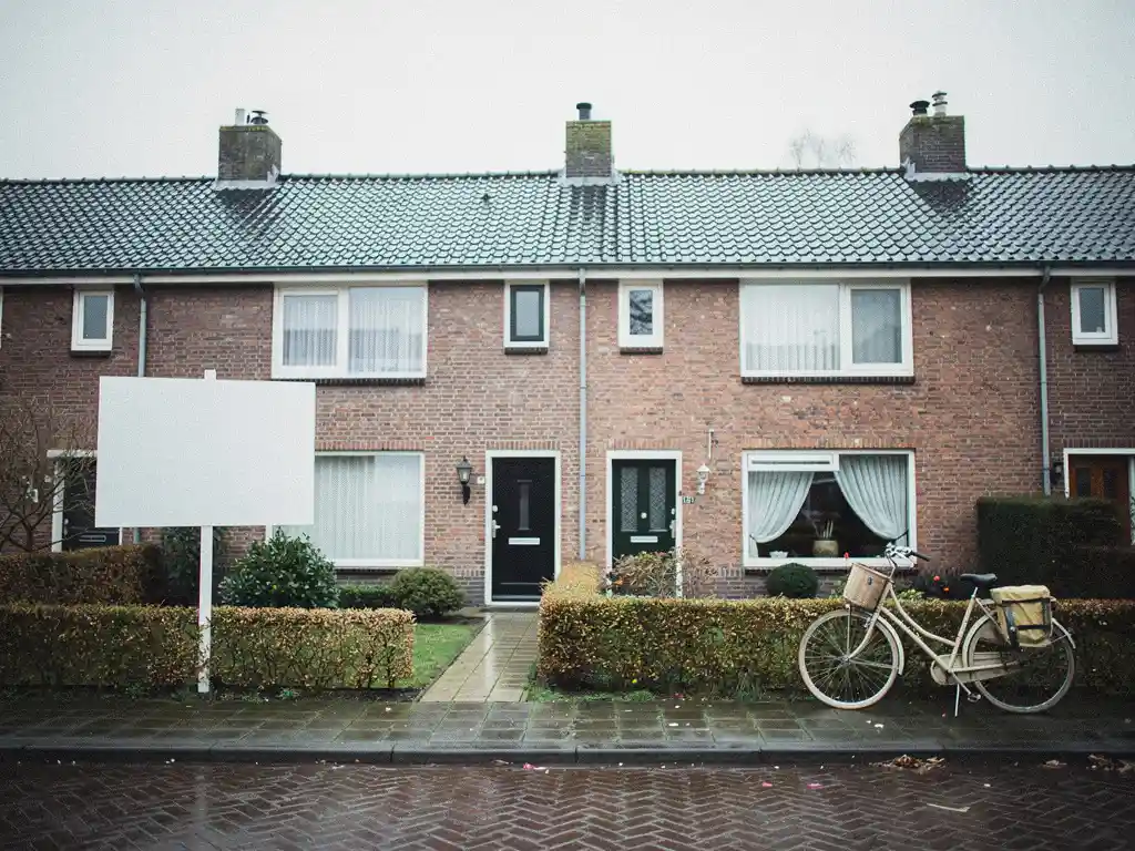Twee identieke bakstenen rijtjeswoningen in Zuid-Holland, één met te koop-bord in voortuin, andere met fiets tegen gevel onder bewolkte lucht.