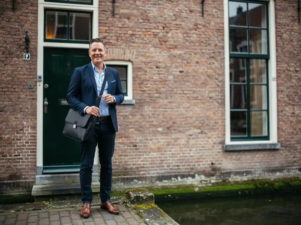 Nederlandse makelaar in casual zakelijke kleding bij de ingang van een 17e-eeuws grachtenpand met trapgevel in Gouda.