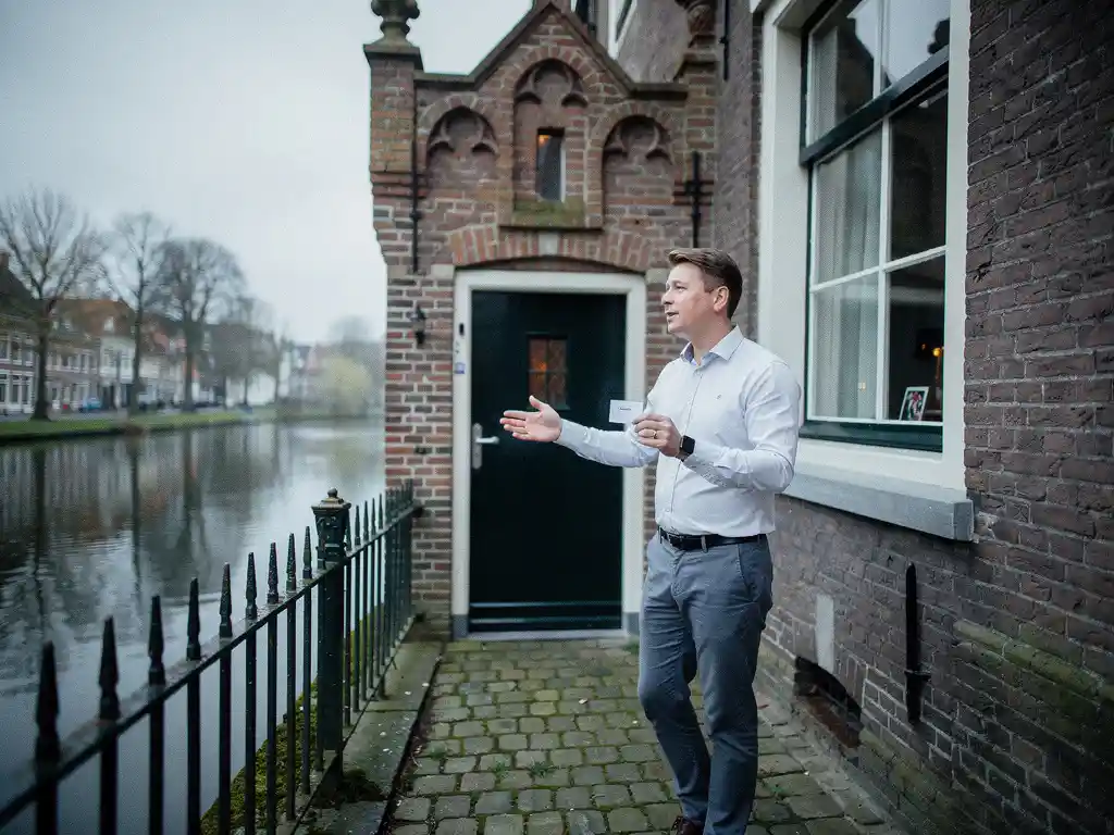 Makelaar in casual businesskleding bij de ingang van een Goudse grachtenpand, wijzend naar de trapgevel met visitekaartje in hand.