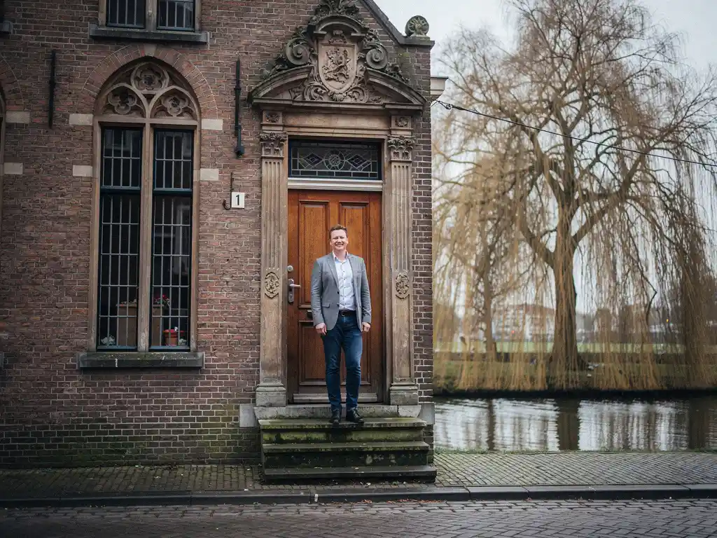Makelaar in zakelijk casual kleding bij de sierlijke houten voordeur van een 17e-eeuws Gouds grachtenpand met trapgevel.
