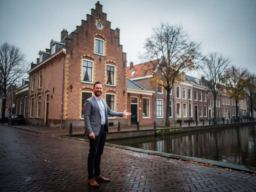 Makelaar in casual zakelijke kleding bij een Hollands grachtenpand met trapgevel in Gouda, herfstige kade met klinkers op bewolkte ochtend.