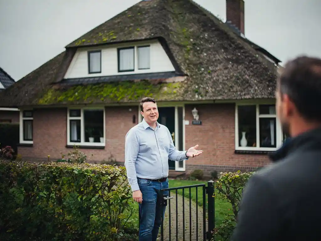 Makelaar in casual zakelijke kleding bij het tuinhek van een jaren 70 bakstenen woning met rieten dak in Reeuwijk.