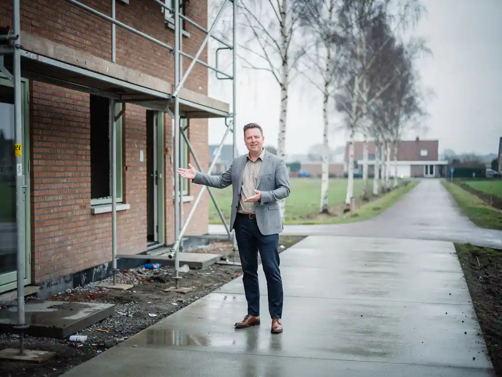 Makelaar in casual zakelijke kleding wijst naar een nieuwbouwwoning van rode baksteen in Gouda, met steigers en bewolkte lucht.