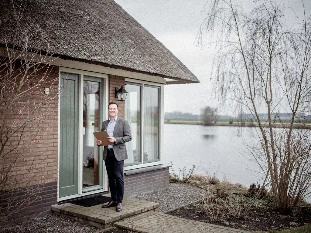 Makelaar met klembord bij de voordeur van een vrijstaande woning met rieten dak in Reeuwijk, polderplas zichtbaar op de achtergrond.
