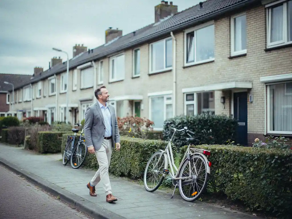 Makelaar wandelt langs jaren '70 rijtjeswoningen in lichtbaksteen op een bewolkte dag in Waddinxveen.