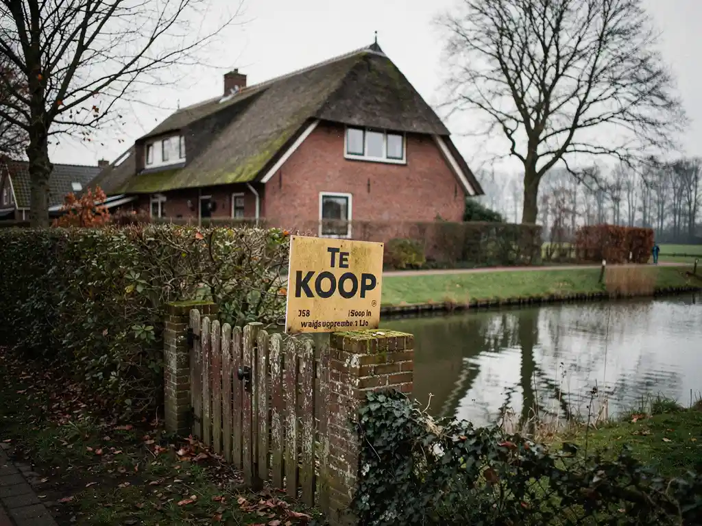 Te koop-bord tegen bakstenen vrijstaande woning met rieten dak in Reeuwijk, omgeven door verwilderde hagen en poldersloot.