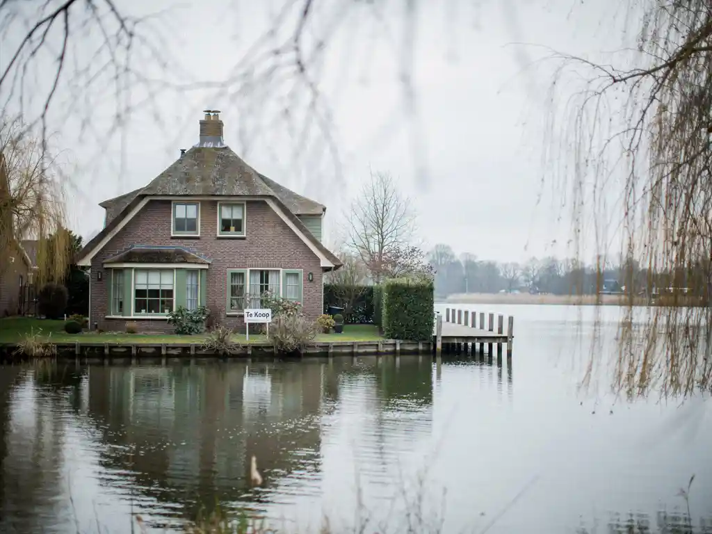 Rieten dak woning te koop aan de Reeuwijkse Plassen, houten steiger over kalm water, kale wilgentakken op bewolkte dag.