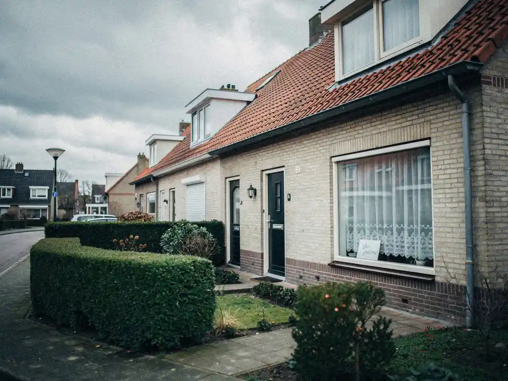 Waddinxveen rijtjeshuis met handgeschreven briefje achter vitrage, lage haag op de voorgrond, bewolkte Nederlandse lucht.