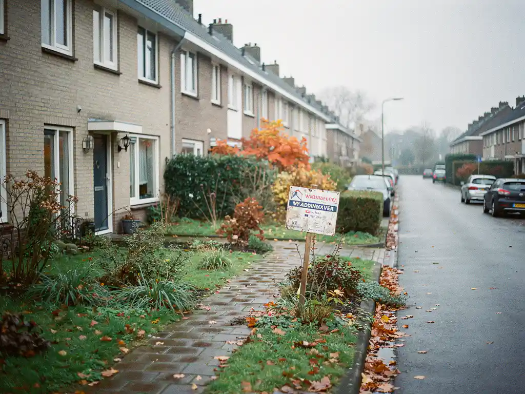 Waddinxveen rijtjeshuis te koop met verweerd bord in verwilderde voortuin, herfstbladeren op nat baksteenpad, bewolkte dag.