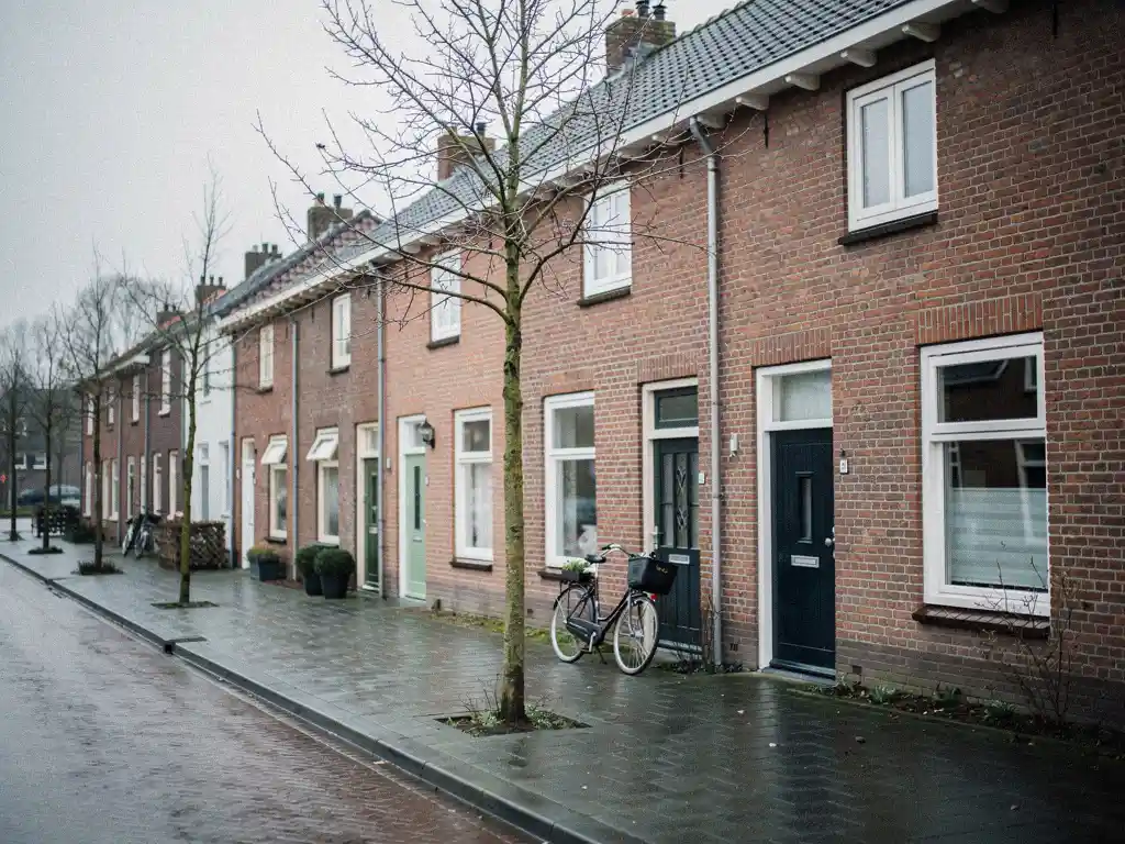 Traditionele rode bakstenen rijtjeshuizen aan een stille straat in Bodegraven, fiets tegen donkere voordeur, kale bomen op nat trottoir.