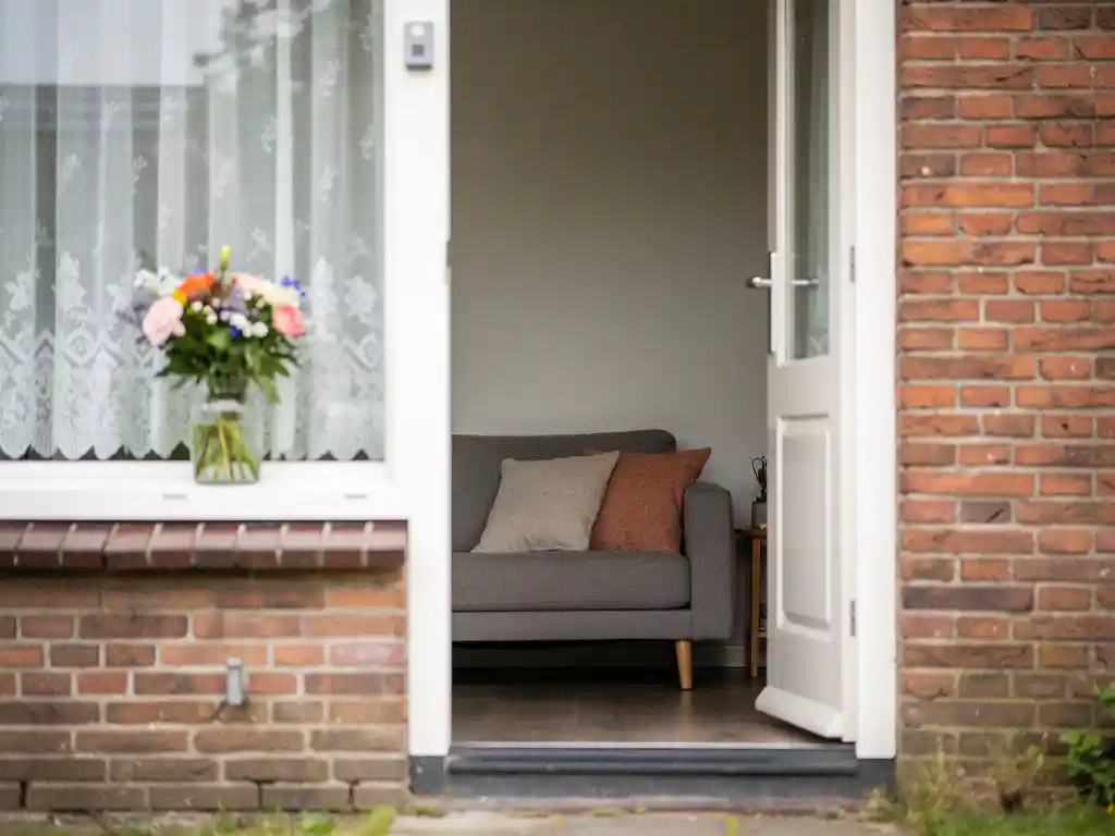 Doorkijkje door open voordeur van rijtjeshuis in Bodegraven naar gezellige woonkamer met linnen kussens en boeket op vensterbank.