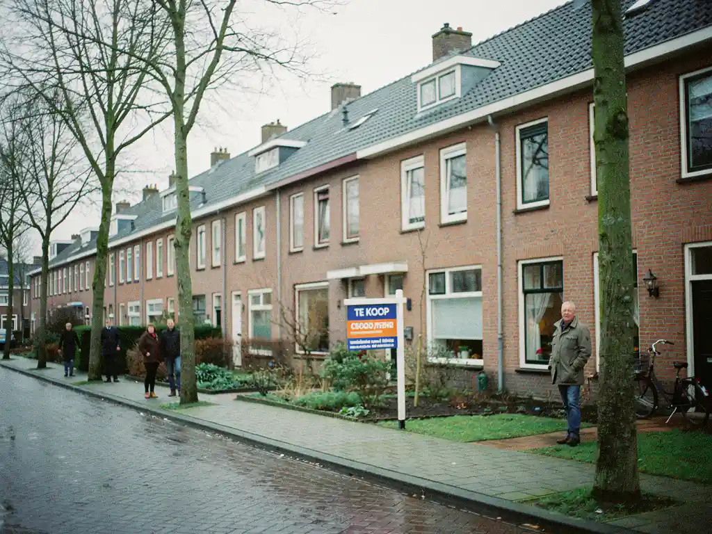 Te Koop-bord voor een rood bakstenen rijtjeshuis in Bodegraven, met meerdere geïnteresseerde kopers op de natte kinderkopjesstraat.